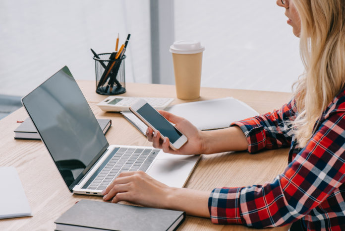 partial view of businesswoman using smartphone and laptop at one time at workplace with papers in newsletter