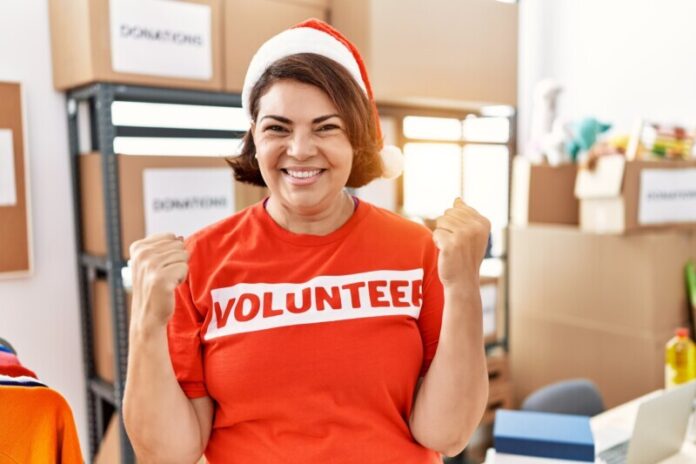 Middle age hispanic woman wearing volunteer t shirt and christmas hat screaming proud, celebrating victory and success very excited with raised arms